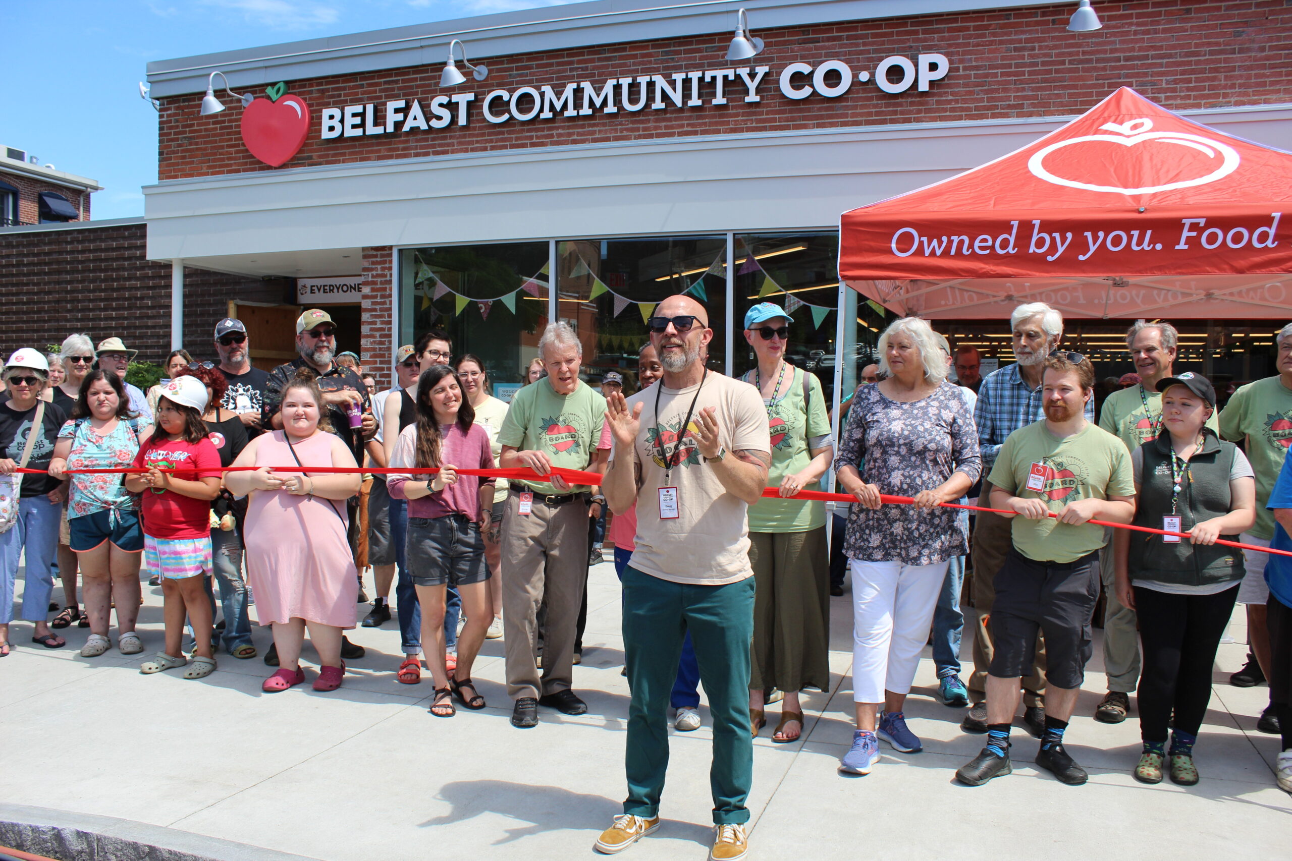 Doug Johnson, General Manager, speaking to crowd of people. People hold a red ribbon behind him in front of the newly renovated Belfast Community Co-op store.