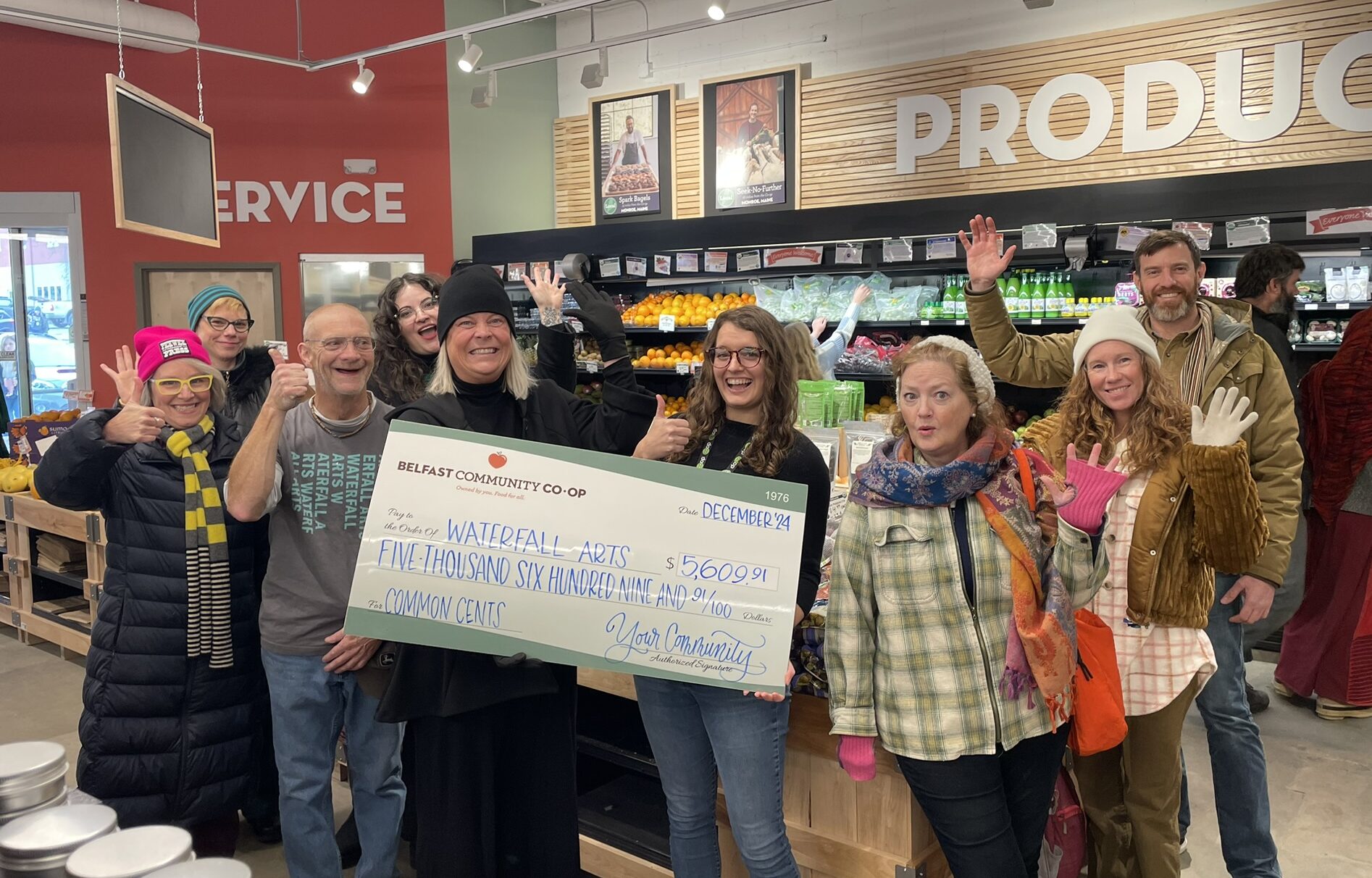 Waterfall Arts Common Cents donation check photo. A group of people holding a giant check with the Belfast Community Co-op Produce department in the background.